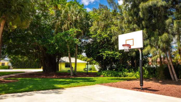 Exterior outdoor space with blue and red hammocks beneath mature trees, green lawn, playground equipment visible, facility building in background