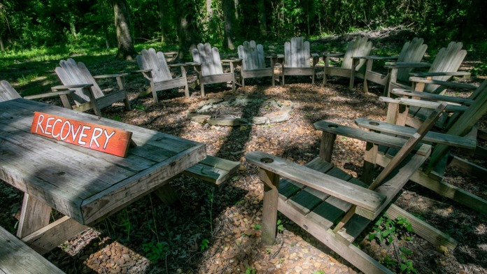 Outdoor space with wooden Adirondack chairs arranged in a circle, marked recovery sign, natural forest setting