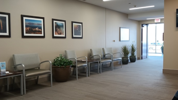 Interior waiting room with comfortable seating, framed artwork on walls, large windows with natural light, and plants