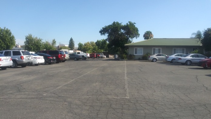 Parking area showing multiple vehicles, facility building with green roof, mature trees, and grounds under clear sky