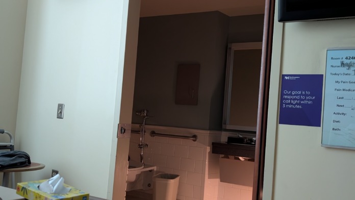 Interior patient bathroom showing toilet, sink, grab bars, wall-mounted fixtures, and access from patient room area with tile flooring