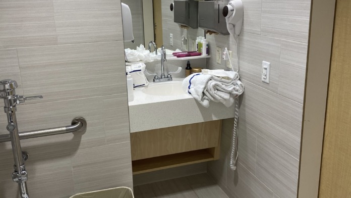Interior bathroom sink area with white and blue-gray tile walls, round basin sink, faucet, paper towel dispenser, hand sanitizer, and mirror.
