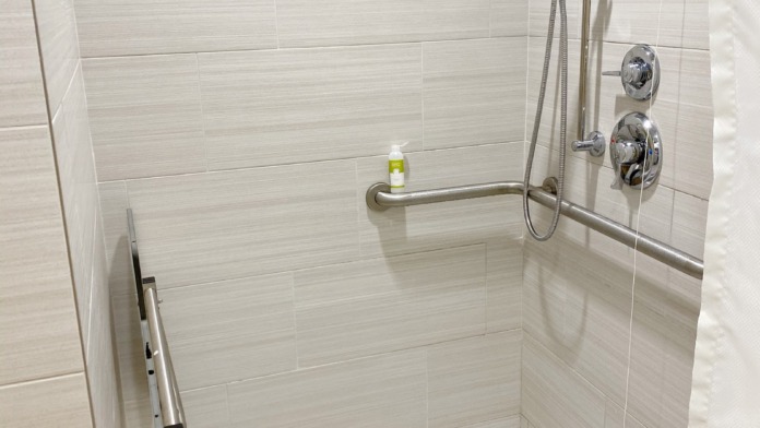 Interior bathroom with white tile walls, decorative blue-gray tile border, walk-in shower with grab bars, modern fixtures, and amenity shelf.