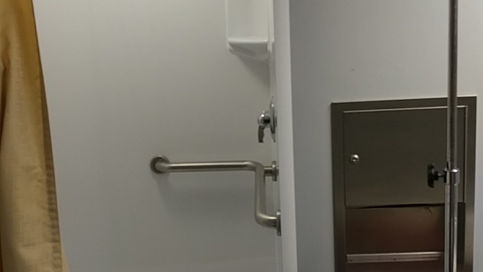 Interior patient bathroom with accessible shower enclosure, grab bars, overhead lighting, and storage shelving