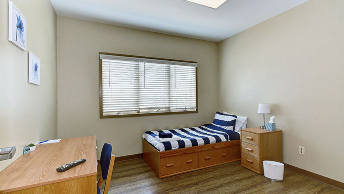 Interior patient bedroom with wooden bed frame with storage, desk, nightstand, window with blinds, and artwork