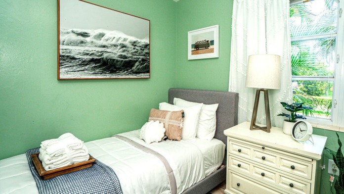 Interior patient bedroom with queen bed, gray upholstered headboard, dresser, nightstand, lamp, and window with garden view
