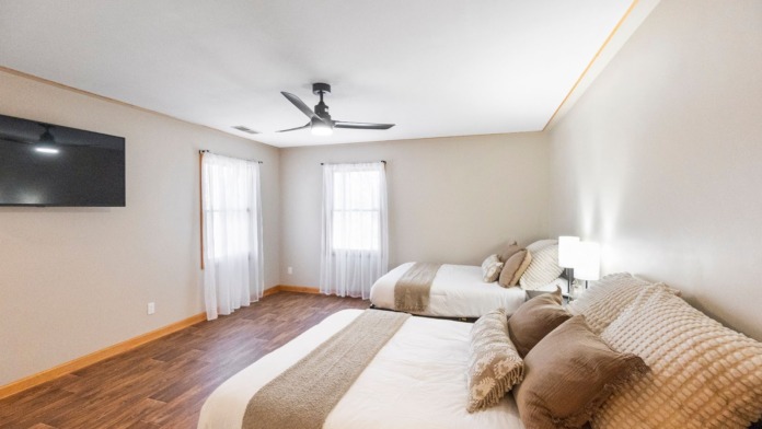 Interior bedroom with two beds with neutral bedding, wall-mounted television, windows with natural light, and hardwood flooring