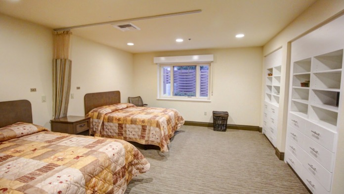Interior patient bedroom with two beds with quilted bedding, nightstands, wooden furniture, window with blinds, and bathroom access
