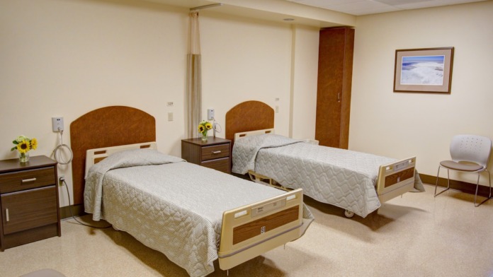 Interior bedroom with two single beds, nightstands with flowers, window with views, medical call button equipment, and comfortable furnishings