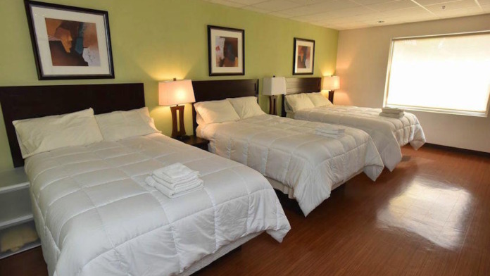 Interior patient bedroom featuring three beds with white linens, nightstands with lamps, framed artwork on walls, and wood flooring