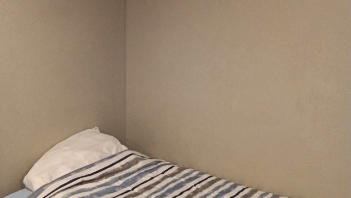 Interior patient bedroom with single bed, striped blue and white bedding, beige walls, and corner placement.