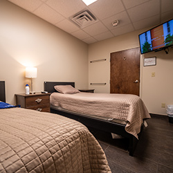 Interior patient bedroom featuring two twin beds with bedding, nightstands, mounted television, and tan walls