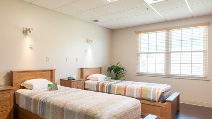 Interior patient bedroom featuring two twin beds with striped quilts, wood nightstand, window with blinds, and plant
