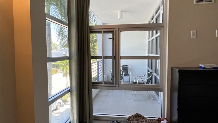 Interior patient bedroom with single bed, dark wood headboard, sconce lighting, artwork, and glass doors opening to water view balcony.