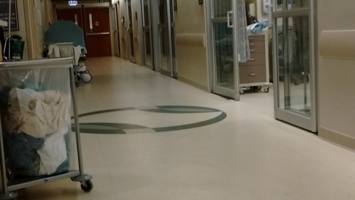 Interior corridor showing numbered patient room doors with glass windows, exit signage, flooring with directional markers, and medical facility equipment