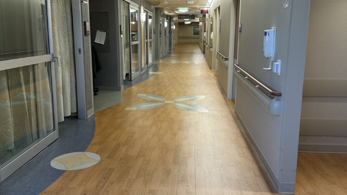 Interior hallway with patient rooms on both sides, wooden flooring, decorative floor patterns, recessed ceiling lights, and medical signage on doors