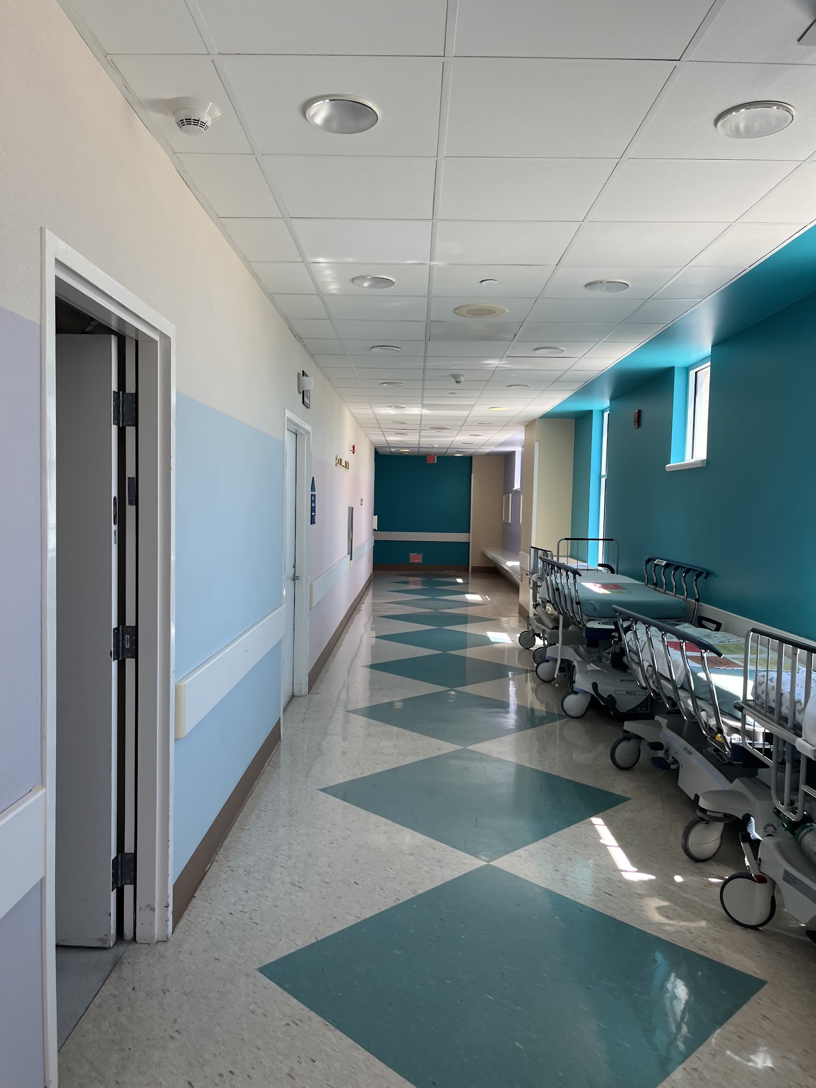 Interior hallway with light blue and teal walls, geometric floor patterns, medical carts and modern ceiling