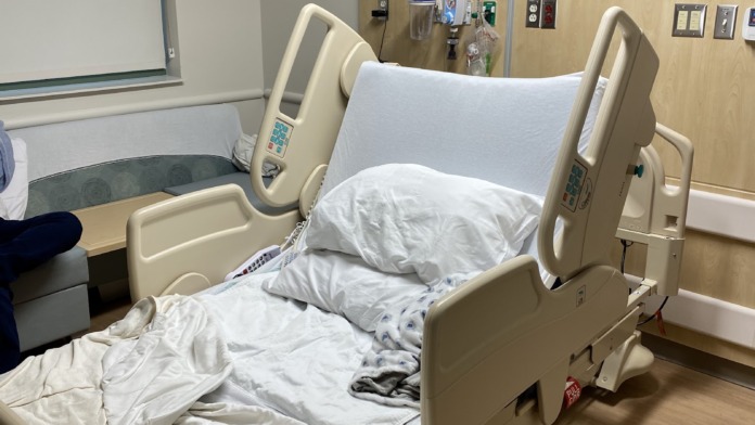 Interior patient bedroom with adjustable medical bed, white bedding, wood accent wall, nature artwork, whiteboard, ceiling-mounted screen, and window.