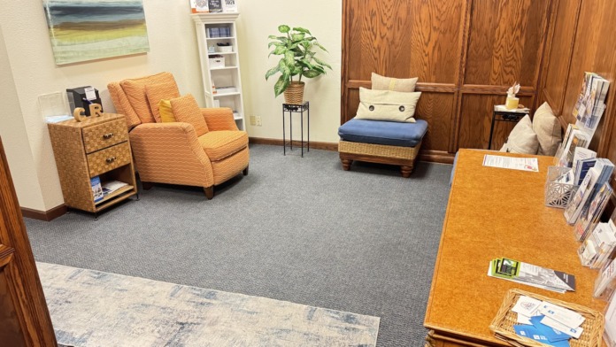 Interior lounge with orange upholstered armchair, wooden furniture, plants, artwork, and informational materials.