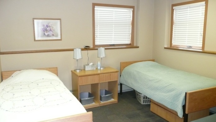 Interior bedroom with two single beds, shared nightstand, windows with blinds, and basic residential furnishings