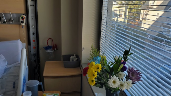 Interior patient room showing desk, chair, fresh flower arrangement, window with blinds, and exterior building view