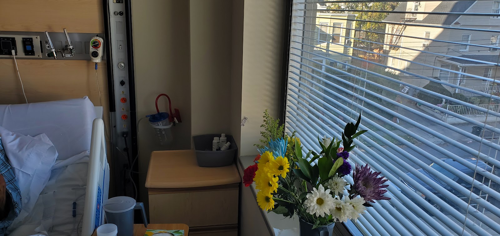 Interior patient room showing desk, chair, fresh flower arrangement, window with blinds, and exterior building view