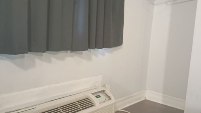 Interior patient bedroom showing single bed with patterned bedspread, window with dark curtains, small desk area, and climate control unit on wall