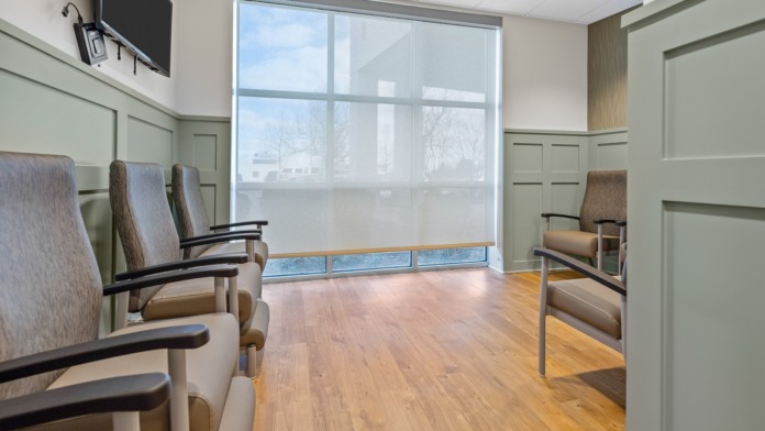 Interior waiting lounge with upholstered seating, large window with exterior views, green wall paneling, and mounted television