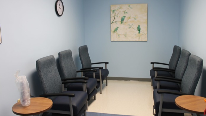 Interior waiting room with rows of gray upholstered chairs, wall clock, decorative bird artwork, and wooden side tables.