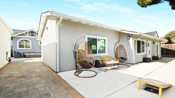 Exterior patio with wicker hanging egg chairs, blue cushions, blue umbrella, and wooden privacy fence