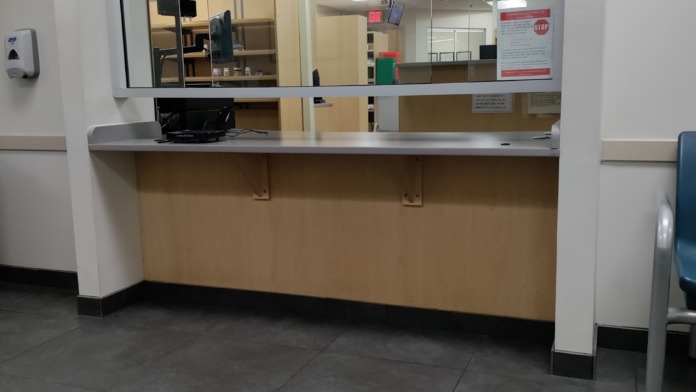 Interior pharmacy service counter with prescription pick-up and drop-off windows, office equipment visible through glass