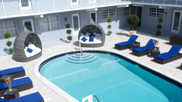 Outdoor pool area featuring resort-style lounge chairs with blue cushions, shade pods, palm landscaping, and modern facility buildings