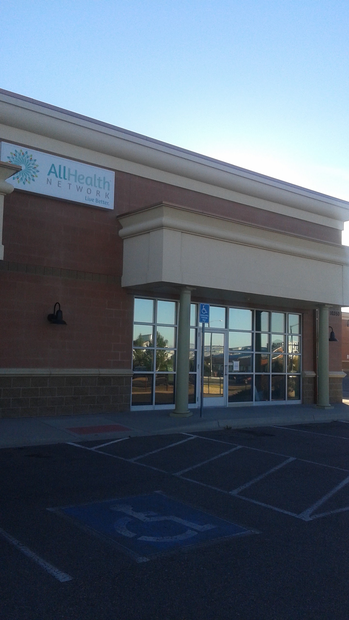 Exterior entrance showing All Health Network signage, glass doors, brick walls, and accessible parking sign