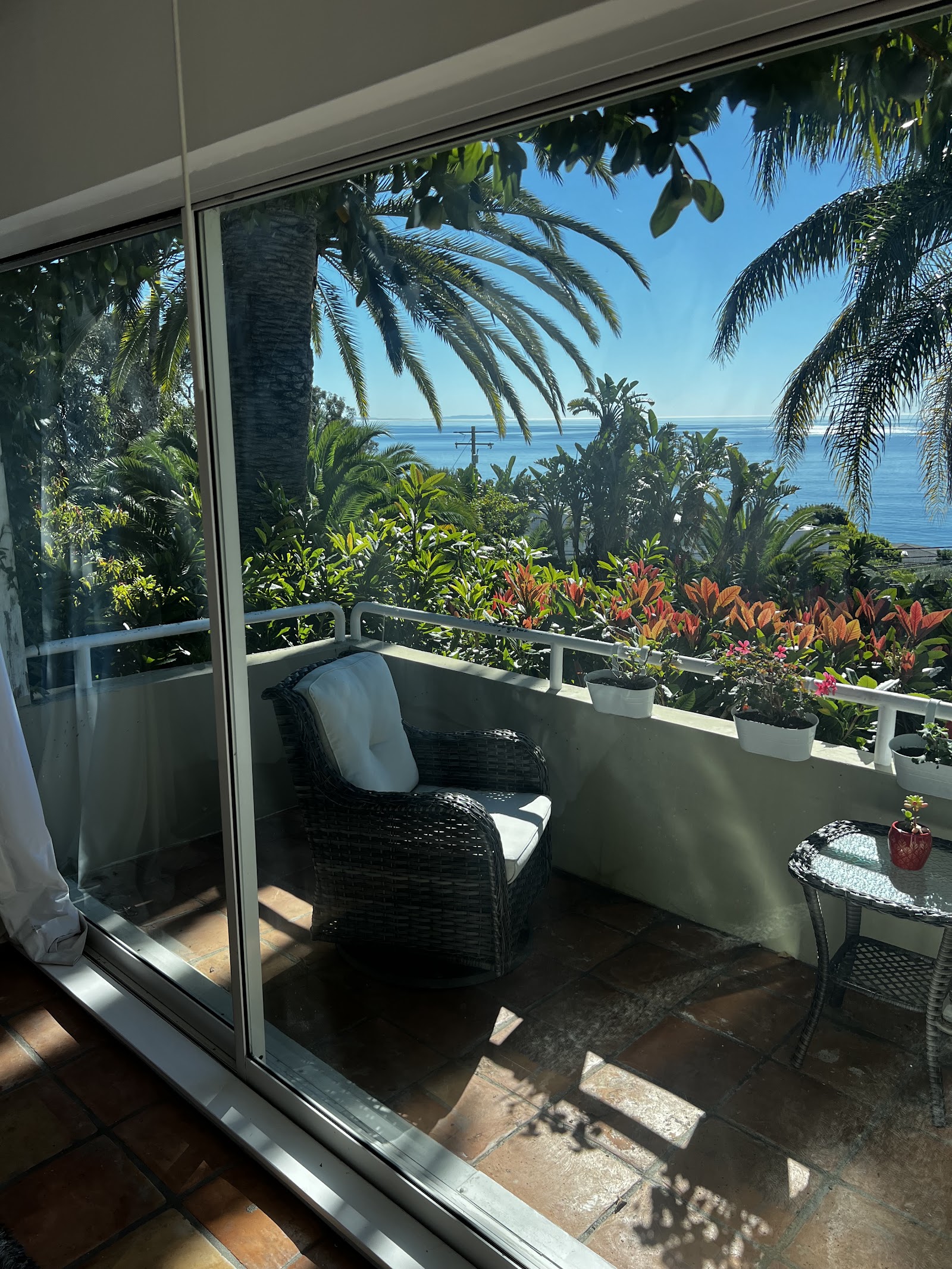 Exterior private balcony with wicker chair, potted flowering plants, and ocean view beyond landscaping
