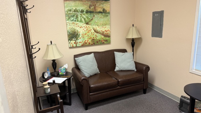 Interior room with brown leather sofa, side table with lamp, framed landscape art, coat rack, and office furnishings.