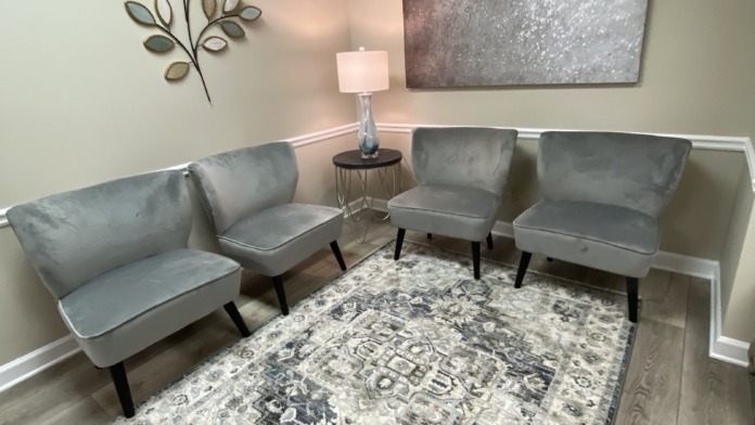 Interior consultation space with four gray upholstered modern chairs, decorative leaf wall art, lamp, and patterned area rug