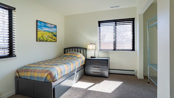 Interior patient bedroom with single bed, black nightstand, patterned bedding, windows with black shutters, and artwork on wall