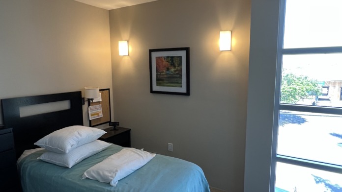 Interior patient bedroom with single bed, green bedding, dark headboard, wall sconces, framed artwork and glass doors to patio.