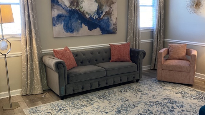 Interior lounge with gray tufted sofa, coral armchair, blue abstract artwork, windows with curtains, and patterned area rug