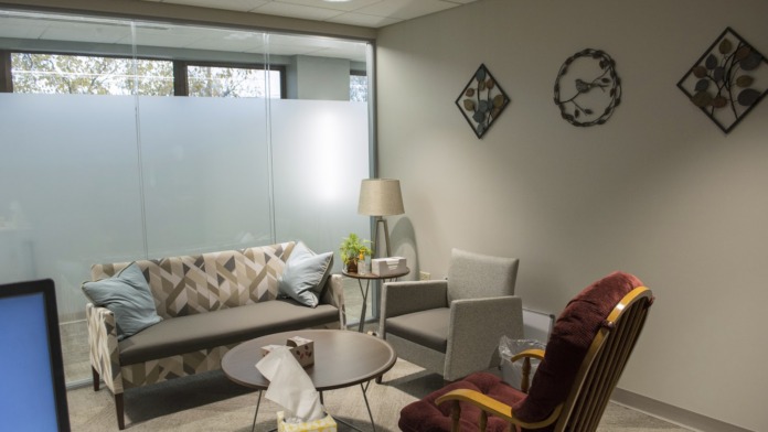 Interior therapy room with gray sofa, patterned pillows, burgundy armchair, side table with lamp, and wall art.