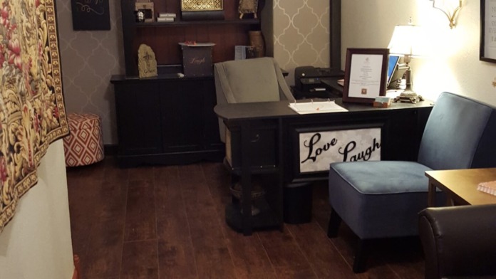 Interior office with wooden desk, shelving unit with decorative items, plants, framed signage, and upholstered seating