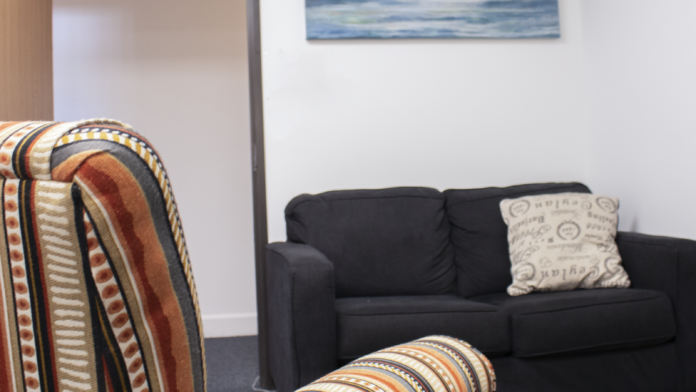 Interior private consultation room with upholstered seating, sofa, decorative pillow, landscape artwork, clock, and peaceful lighting for individual sessions