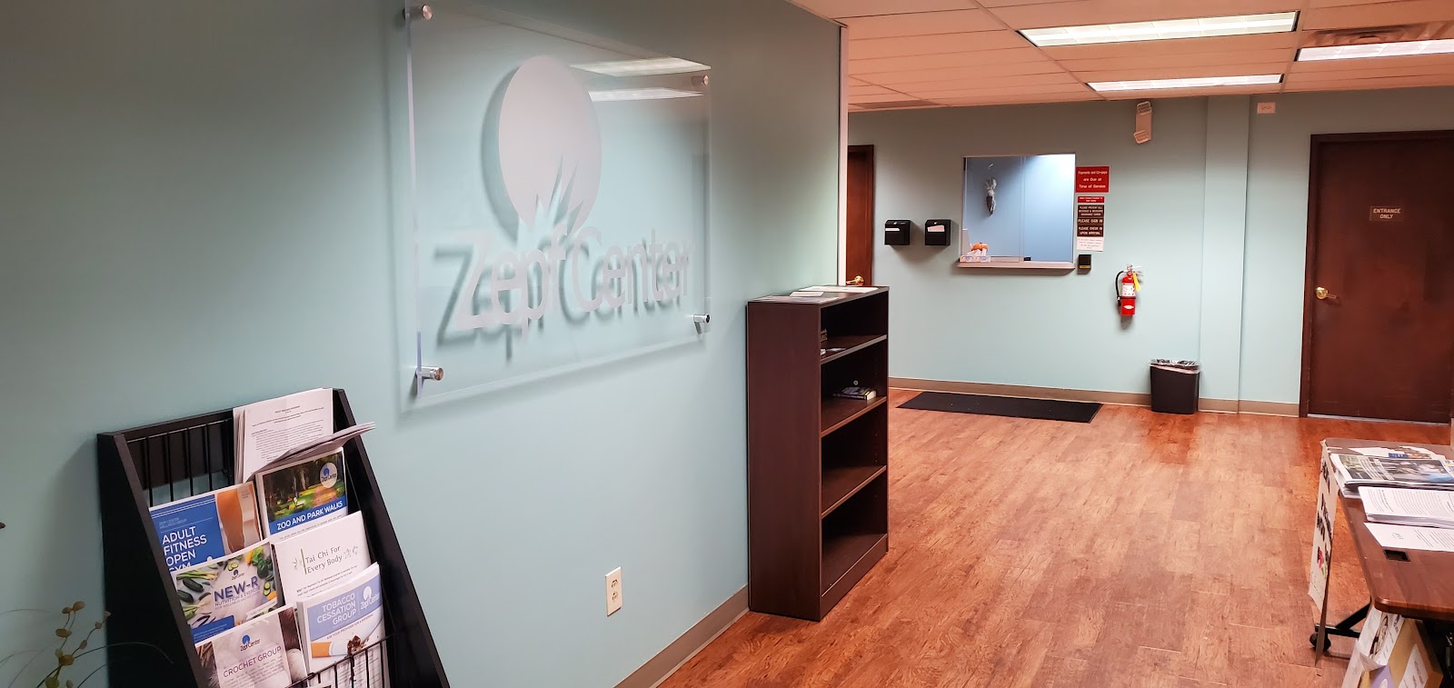 Interior reception area with Zepf Center logo on wall, wooden flooring, shelving unit, and administrative workspace with signage and documents