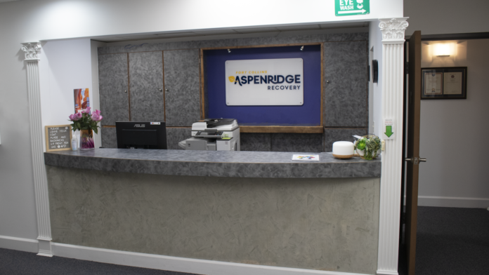 Interior administrative reception desk with signage displaying AspenRidge Recovery logo, computer, office supplies, and professional check-in counter