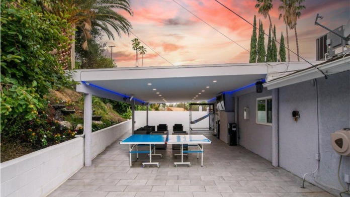 Outdoor covered recreation space with ping pong table, blue awning, hillside setting, and cypress trees visible.