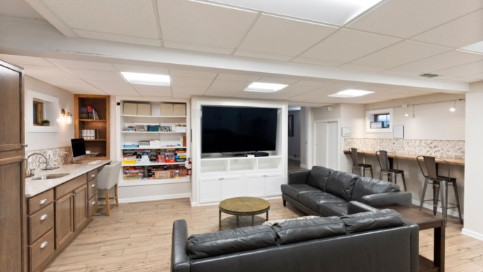 Interior recreational space with multiple black leather couches, entertainment center with large television, shelving with games and books, and bar seating area