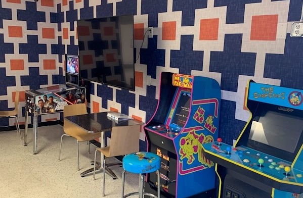Interior game room with arcade machines, geometric patterned wall mural in blue and orange tones, and entertainment seating