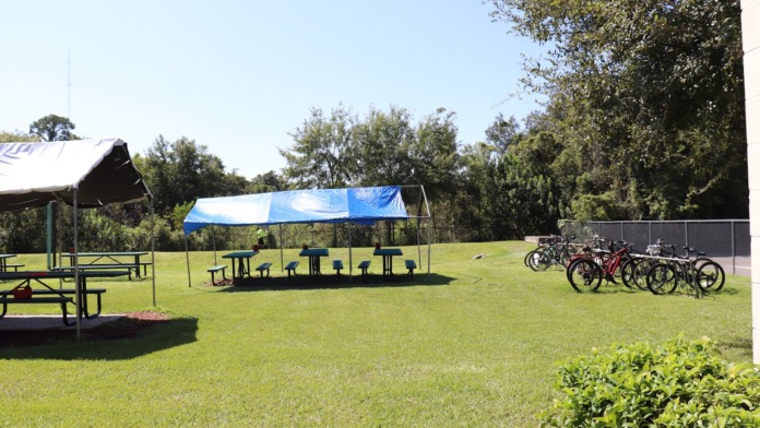 Outdoor recreational space with picnic tables, canopies, bikes, and green lawn surrounded by trees