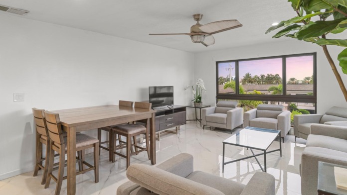 Interior common lounge with wooden dining table, comfortable seating, television, large windows overlooking palm trees, and natural lighting