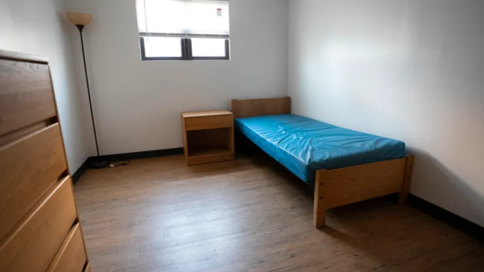 Interior bedroom with single bed, blue mattress, nightstand, lamp, and hardwood flooring in residential facility